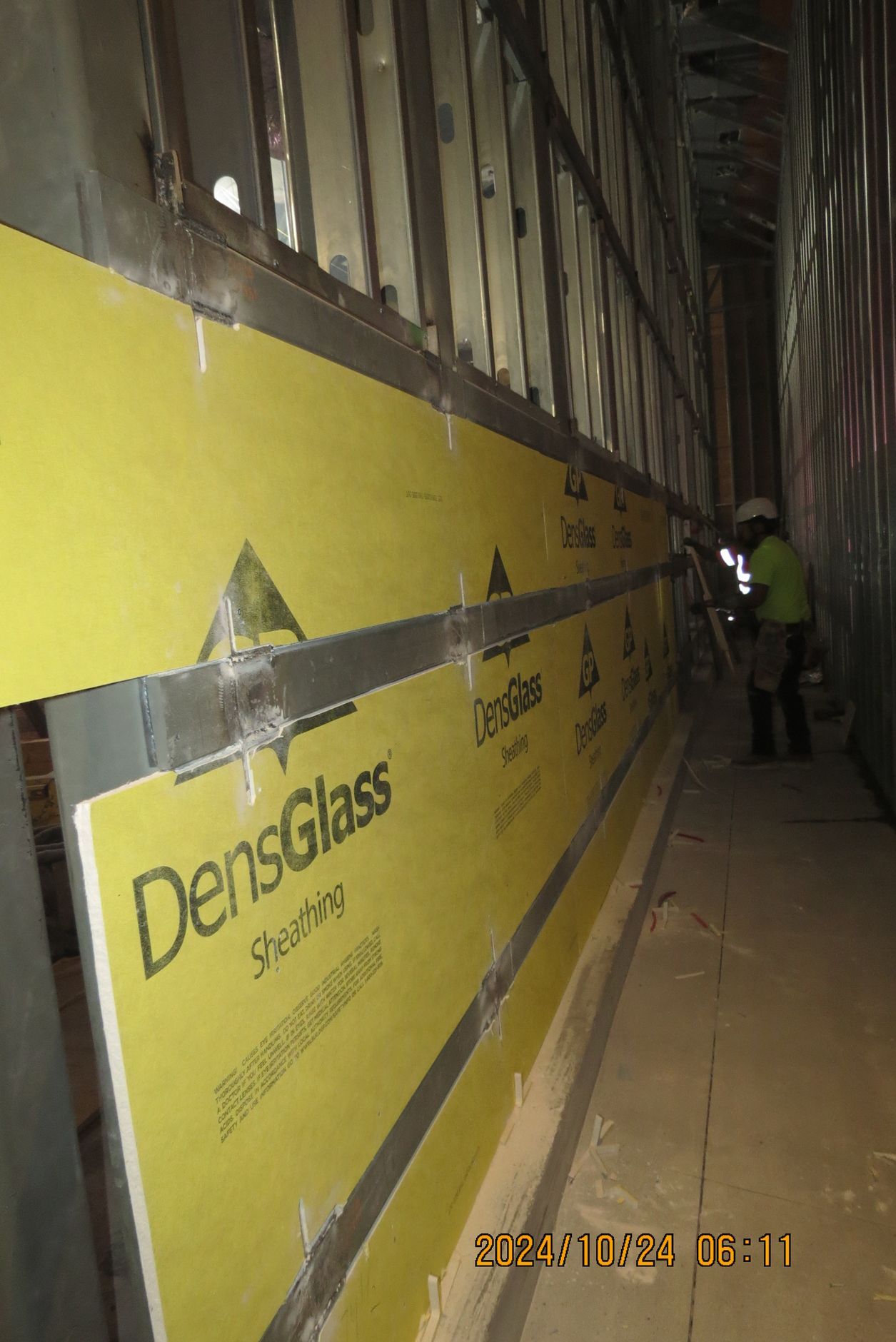 Construction worker installing yellow DensGlass sheathing on a metal stud wall inside a building.