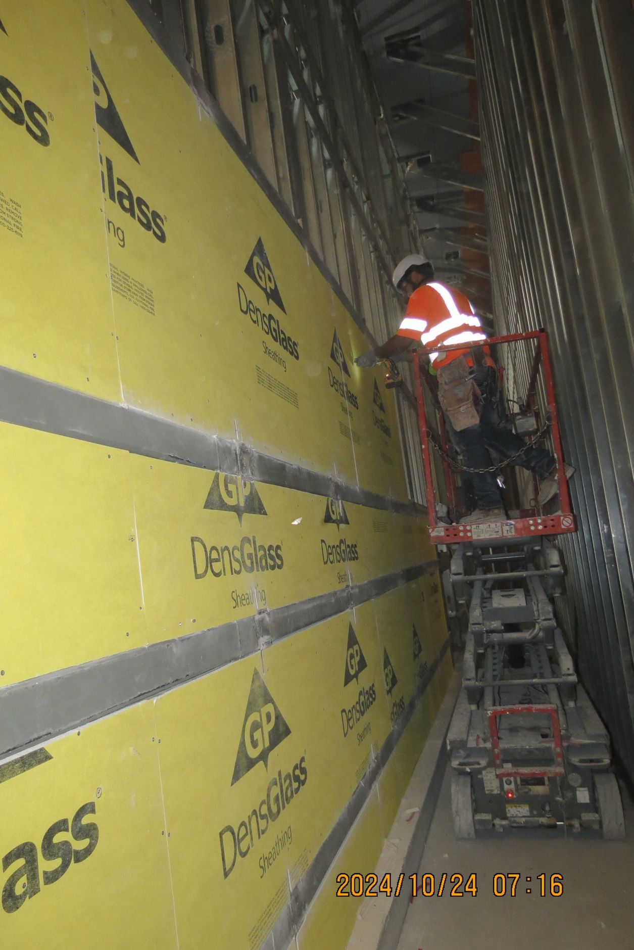 Construction worker on lift installing yellow insulation boards within a metal-framed wall.