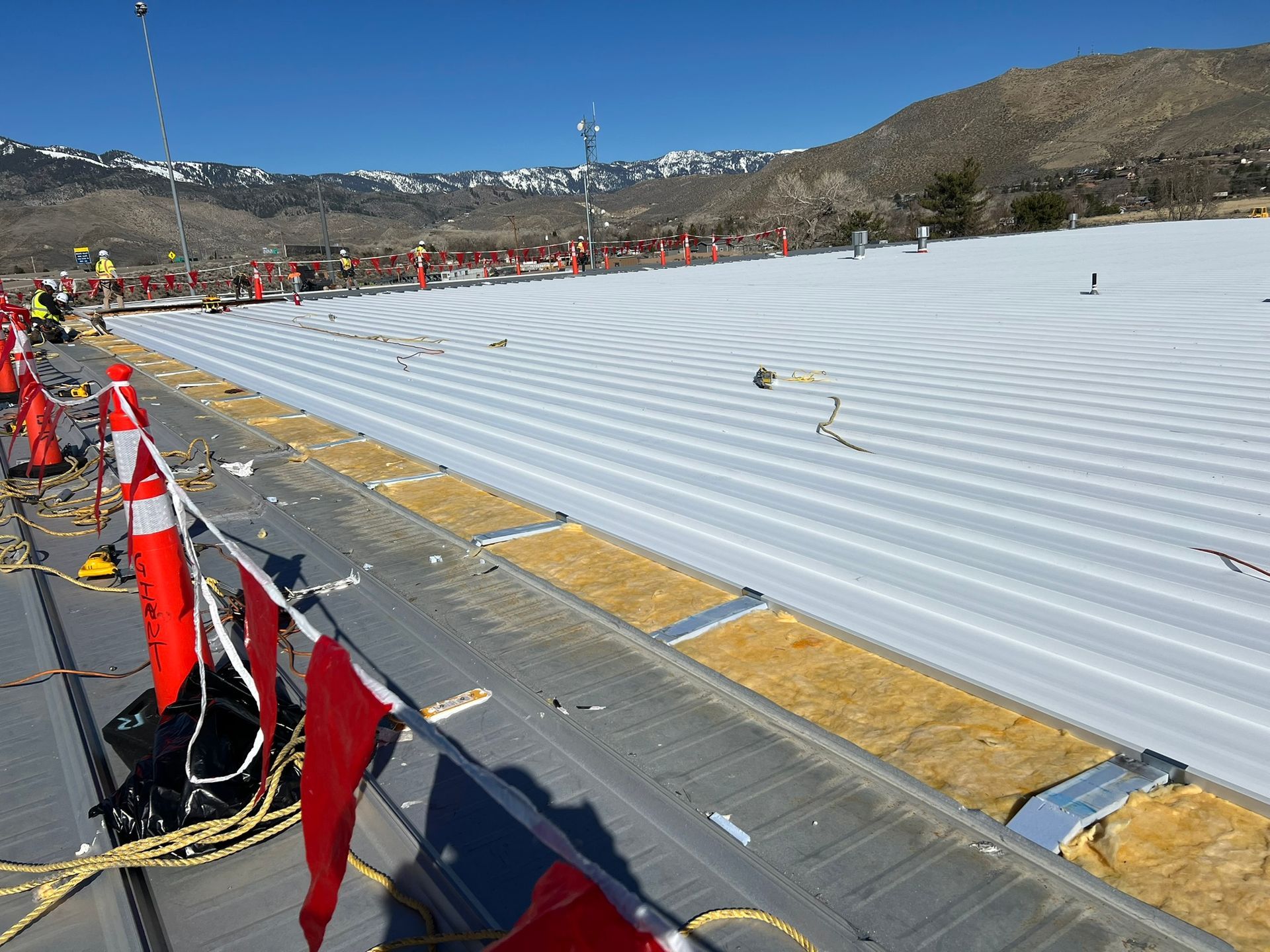 Construction site: large roof covered with white material, red flags, cones, workers, and a mountain backdrop.
