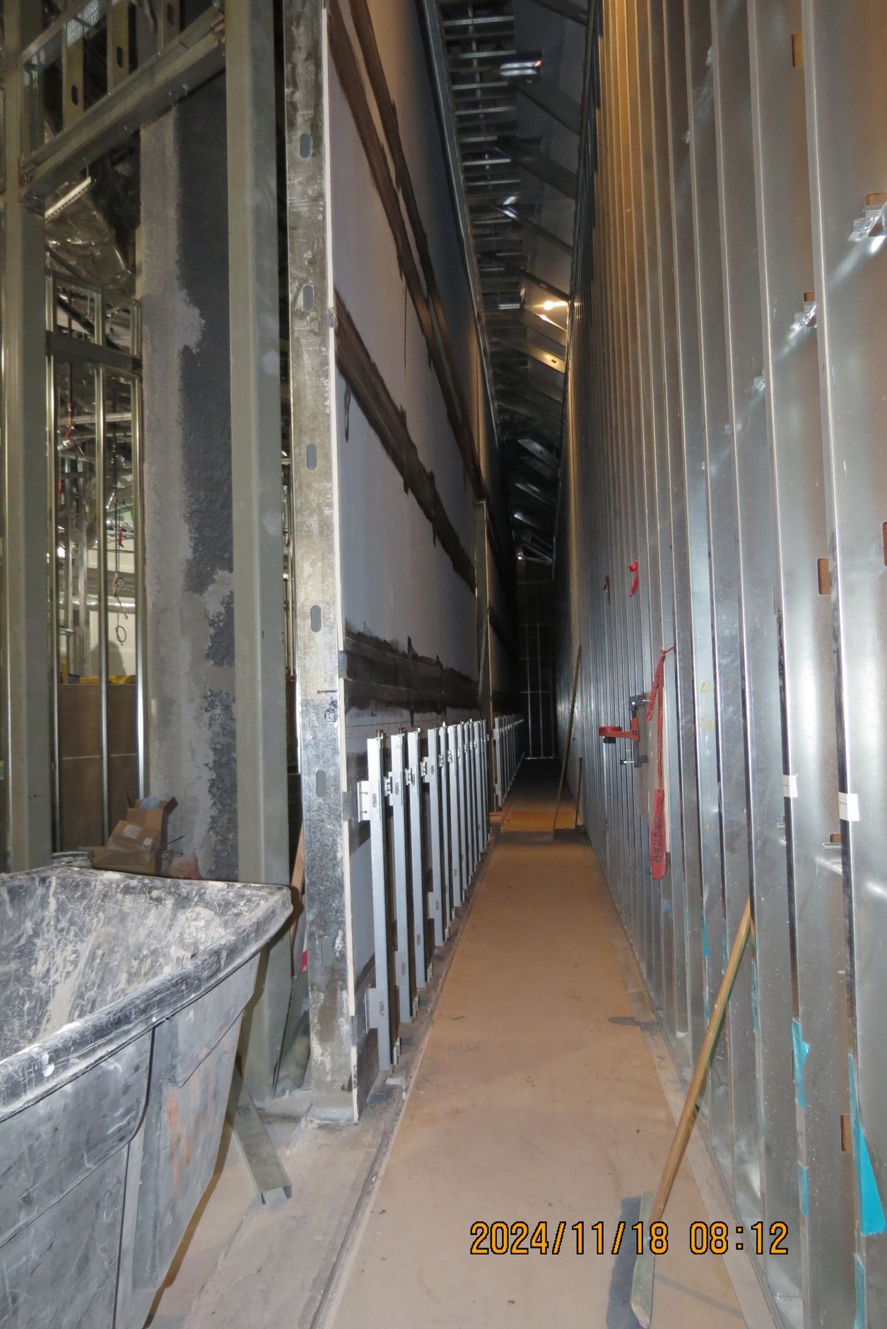 Narrow construction hallway framed in metal studs, with a wheelbarrow of materials on the left.