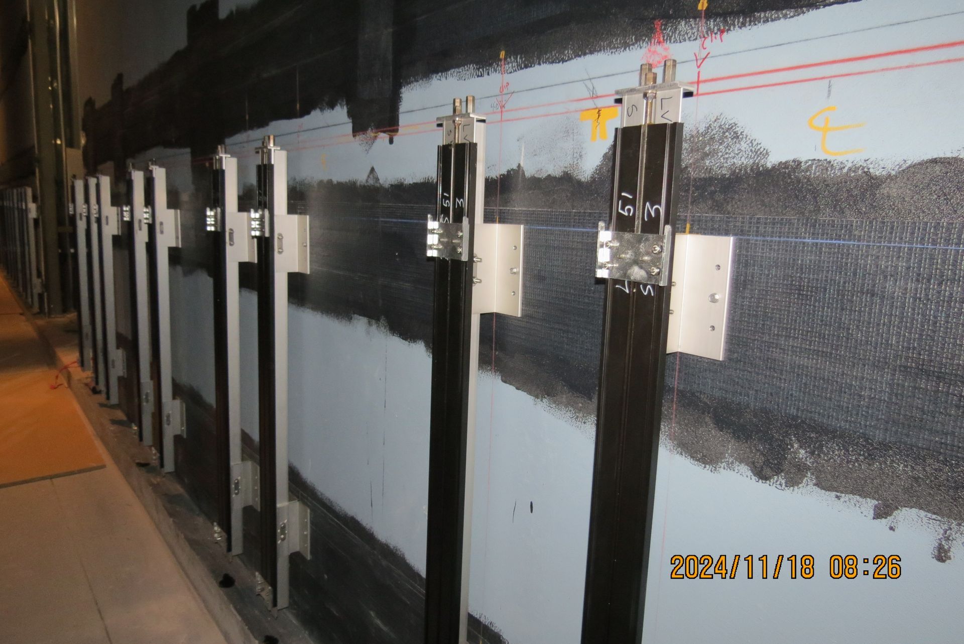 Dark cables secured to a wall by metal brackets. The wall is gray with black markings.