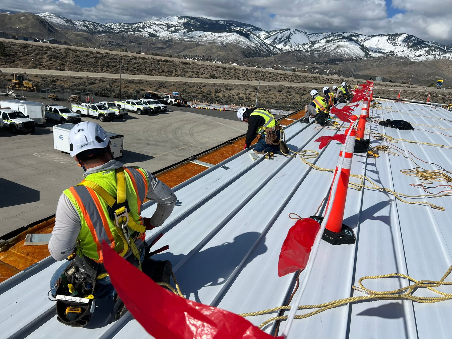 Construction workers in safety gear on a metal roof, with mountains in background.