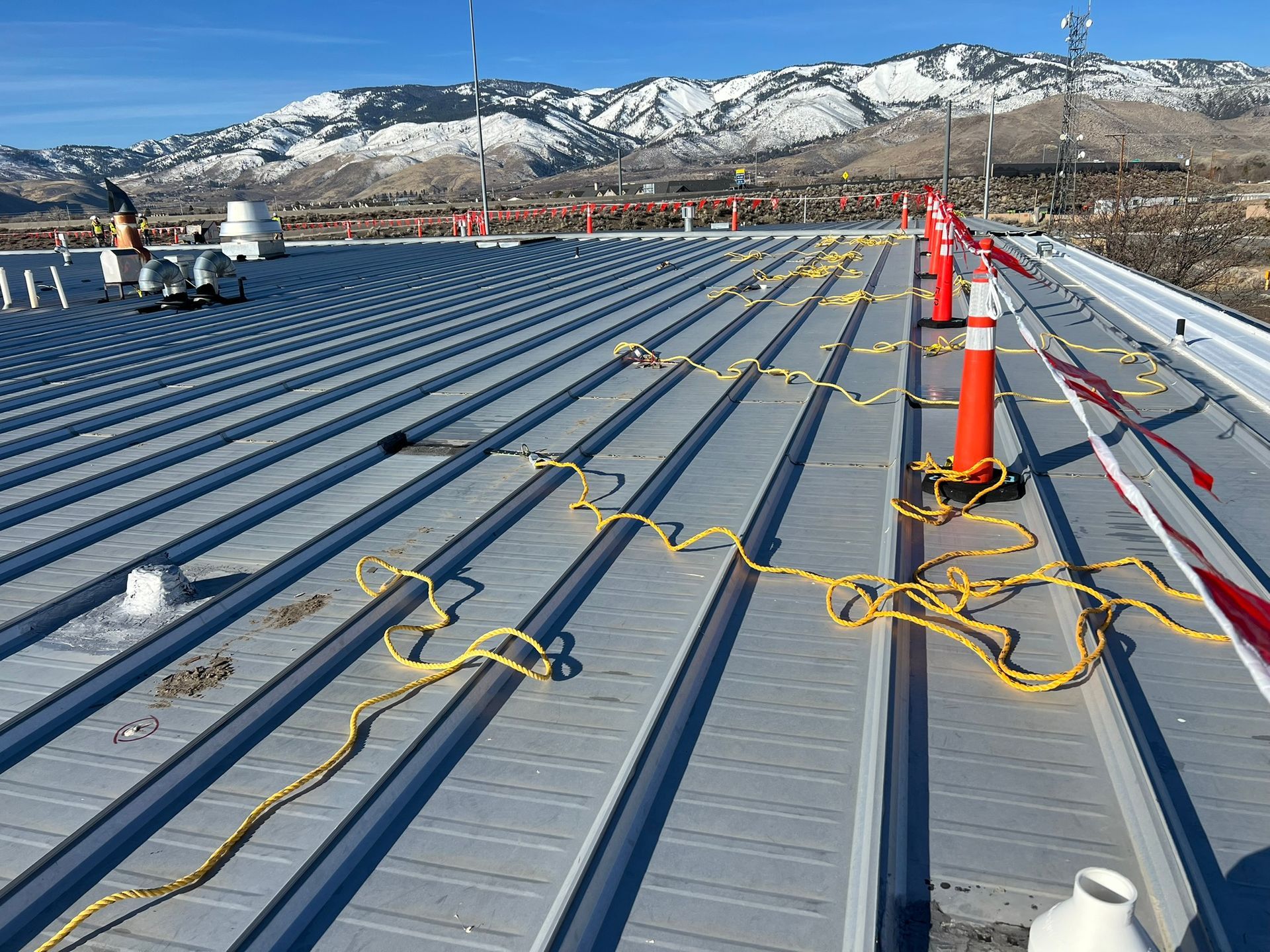 Workers on a metal roof with safety cones and yellow cords. Mountains and blue sky in the background.