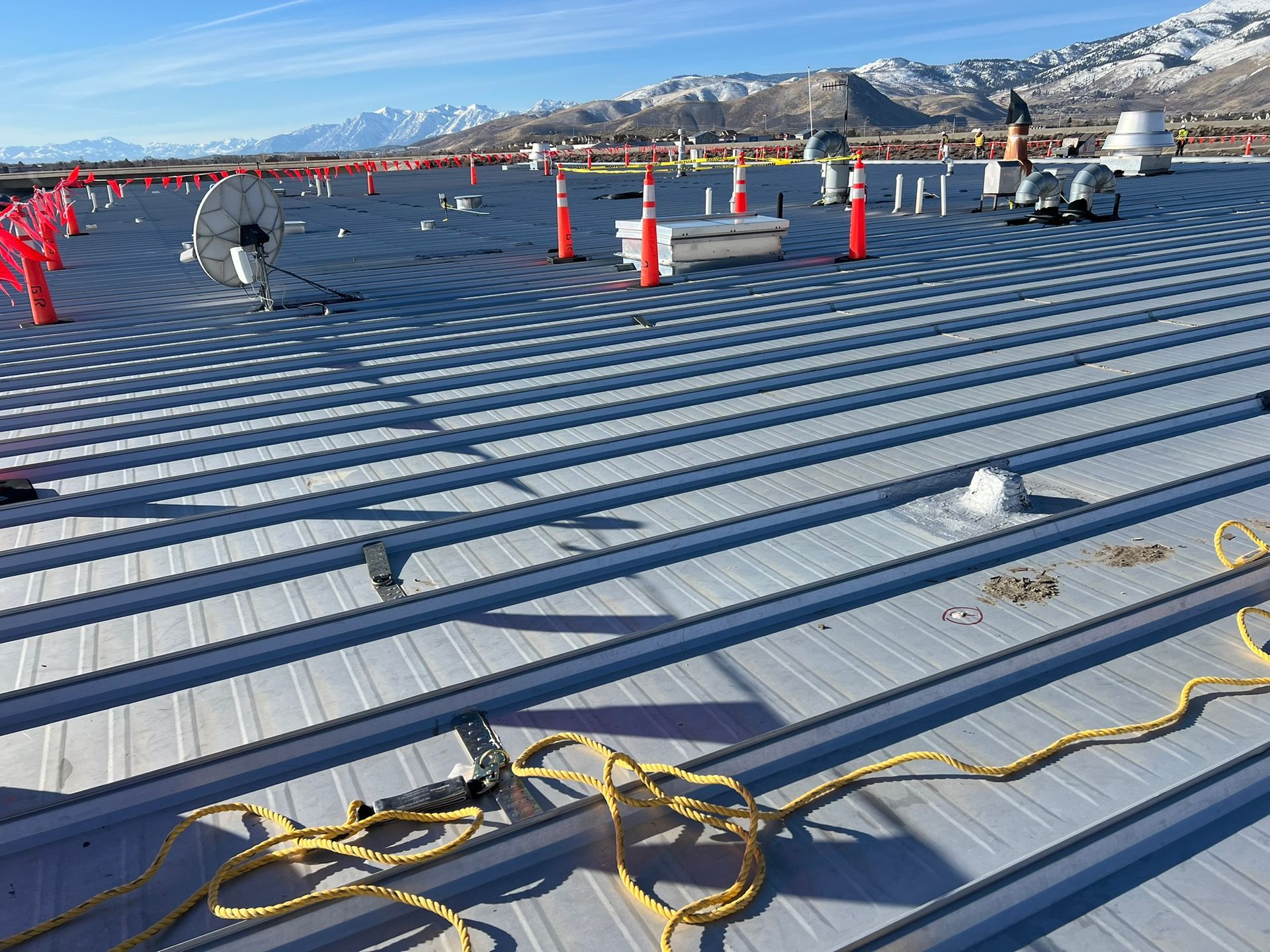 Metal roof construction site with orange safety cones, power cords, and mountain backdrop.