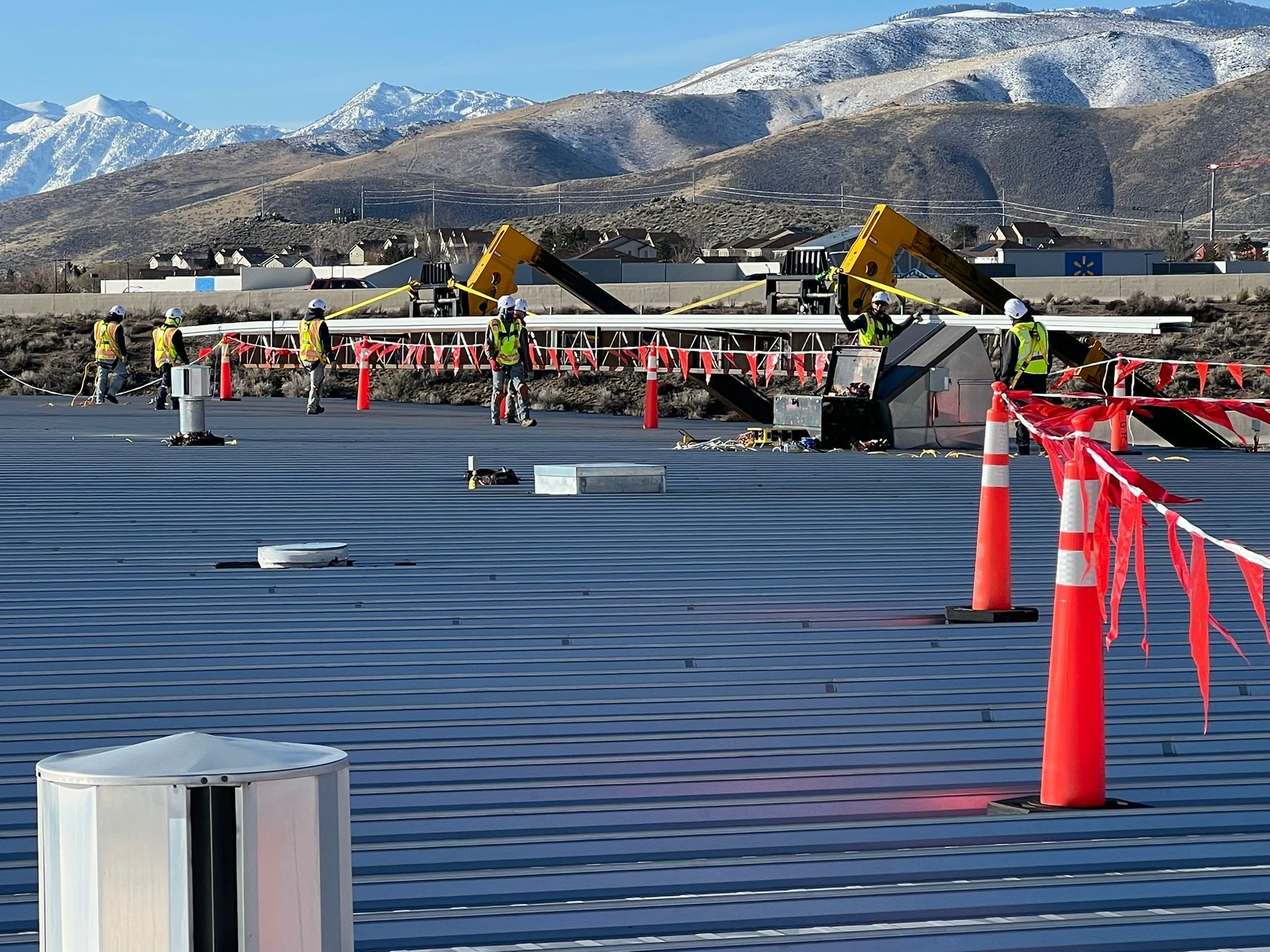 Construction workers on a flat roof, orange safety cones, with mountains in the background under a blue sky.