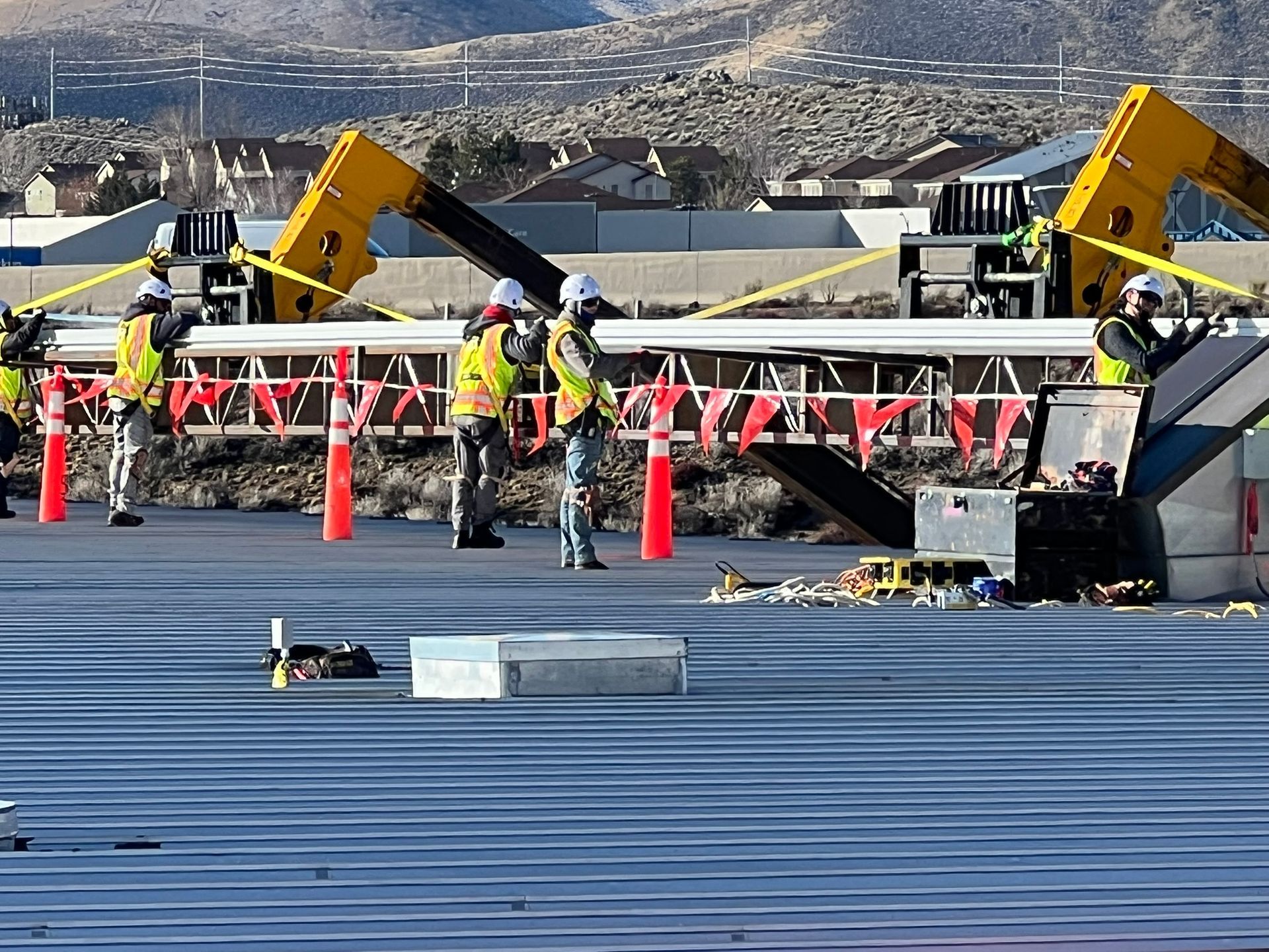 Construction workers placing a concrete beam on a bridge; bright vests, safety helmets, machinery, and a suburban backdrop.