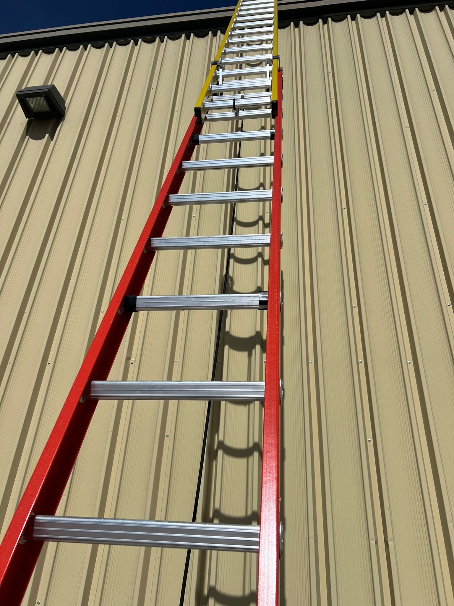 Red and yellow ladder leaning against a beige corrugated metal wall, ascending towards the top.