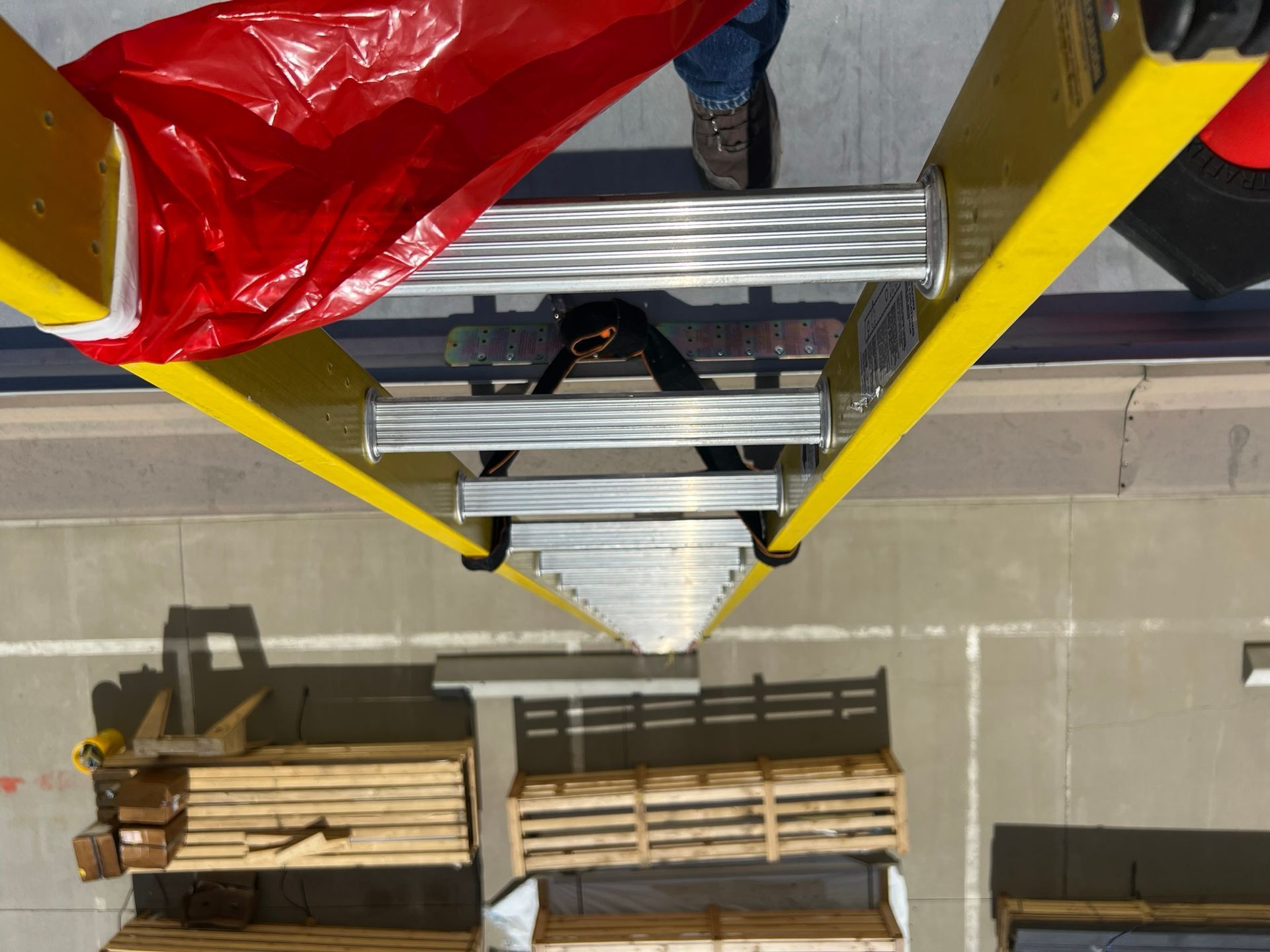Yellow ladder angled down, with a person's legs visible near top. Pallets and flooring are visible below.
