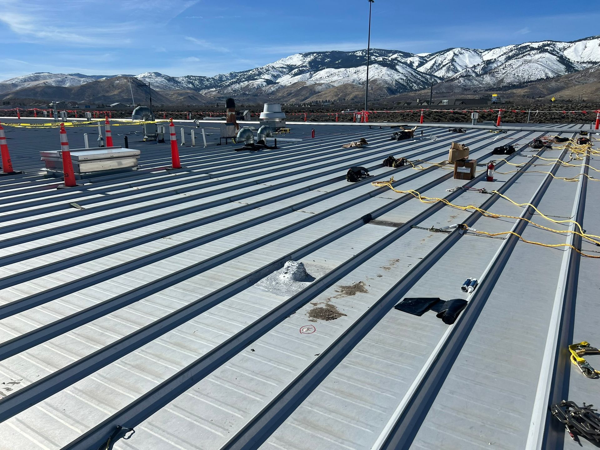 Metal roof under construction with safety cones, equipment, and a snowy mountain backdrop.
