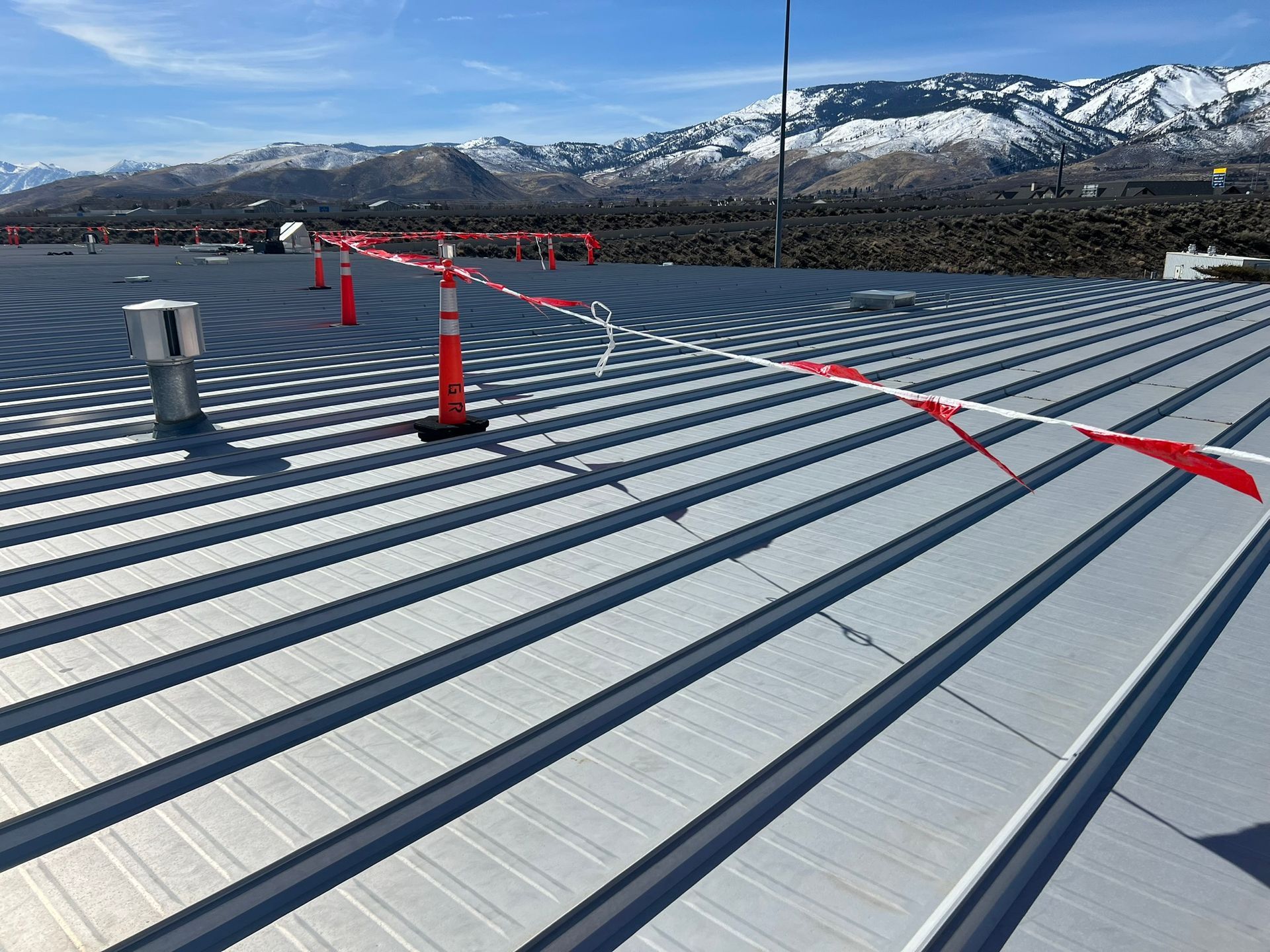 Metal roof with orange safety cones and red/white tape, snowy mountains in the background.