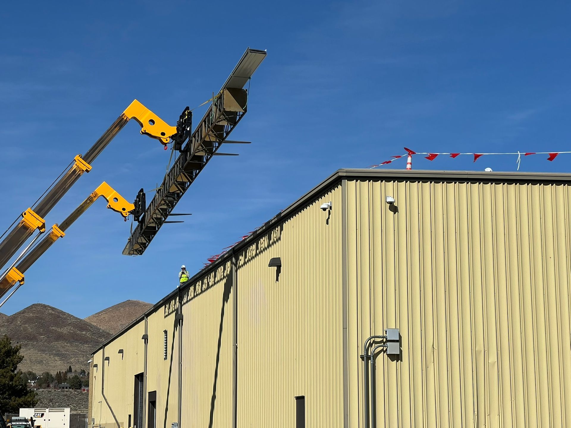 A construction crane lifts a metal structure onto the roof of a beige building on a sunny day.