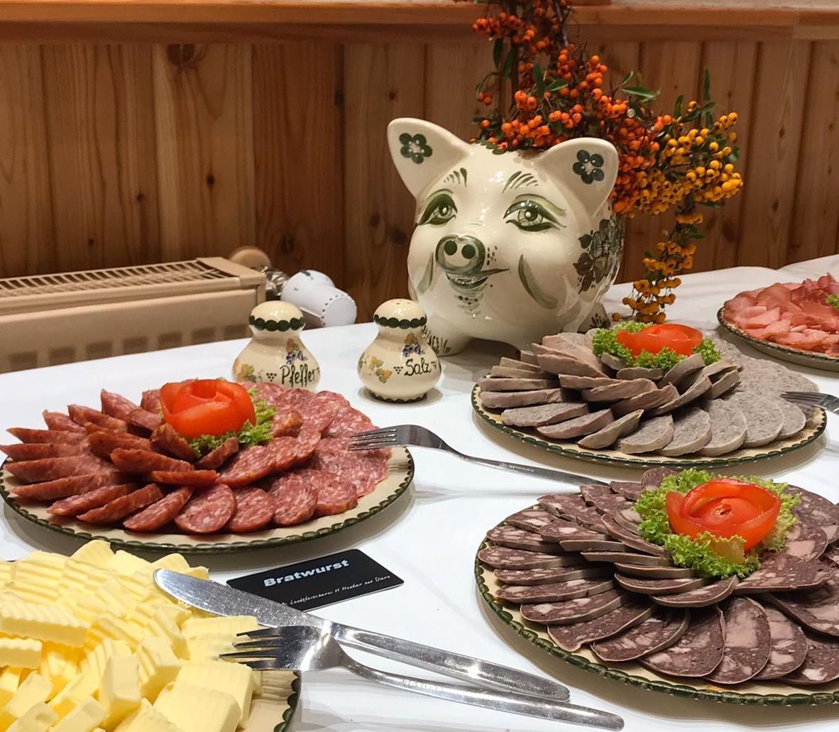 Buffet mit Wurstscheiben, Butter und einer Dekoration in Schweineform. Gedeckter Tisch mit auf Tellern angerichteten Speisen und herbstlichen Blumen.