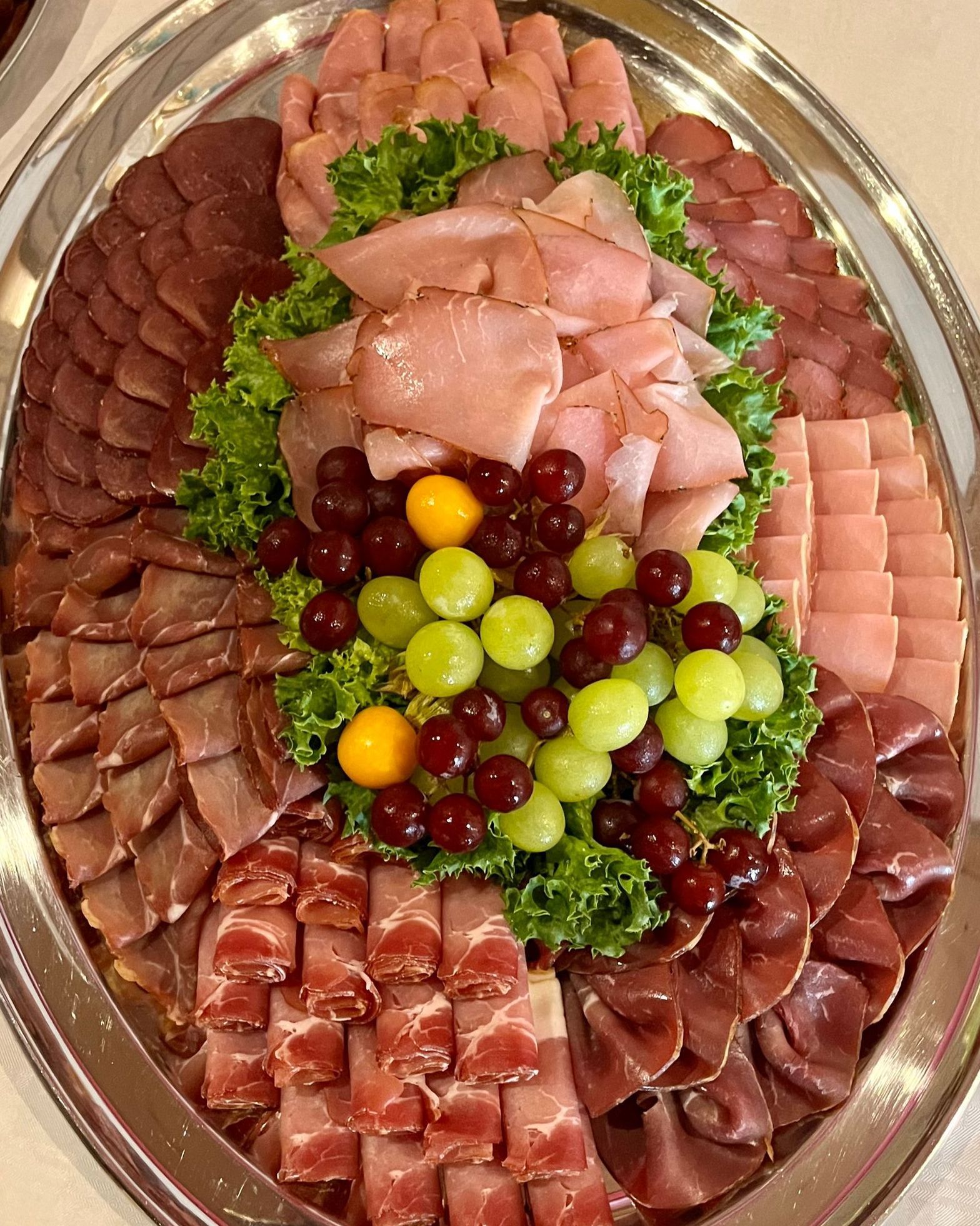 Ovale Platte mit einer Auswahl an Aufschnitt, Trauben und Salat; angerichtet auf einer Silberplatte.