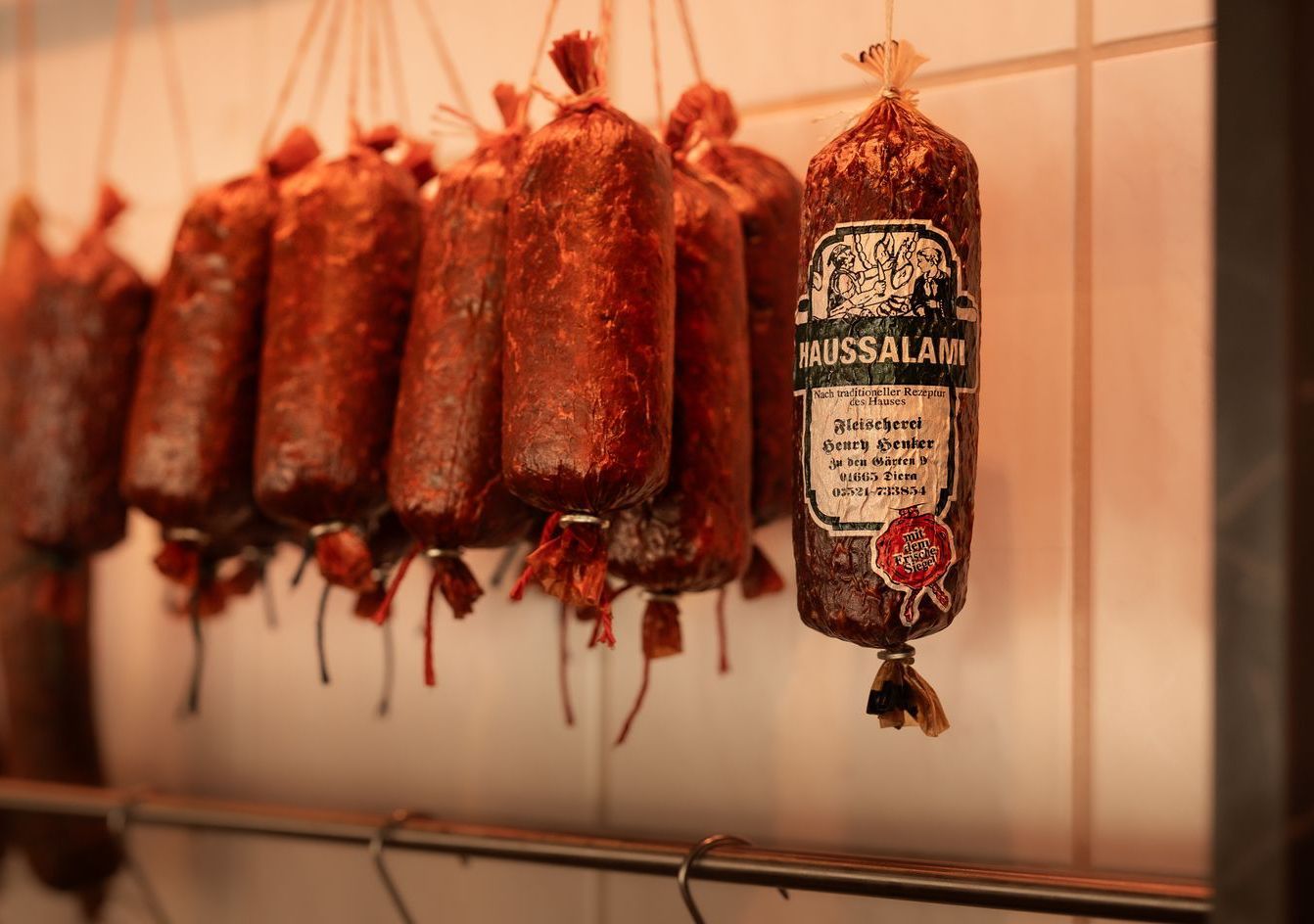 Reihen dunkler, getrockneter Würste hängen an Schnüren in einer Metzgerei. Eine Wurst hat ein Etikett.