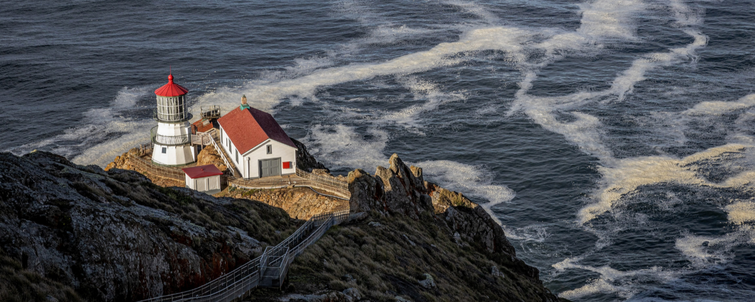 Lighthouse with a red roof and tower on a rocky cliff overlooking turbulent ocean waters.
