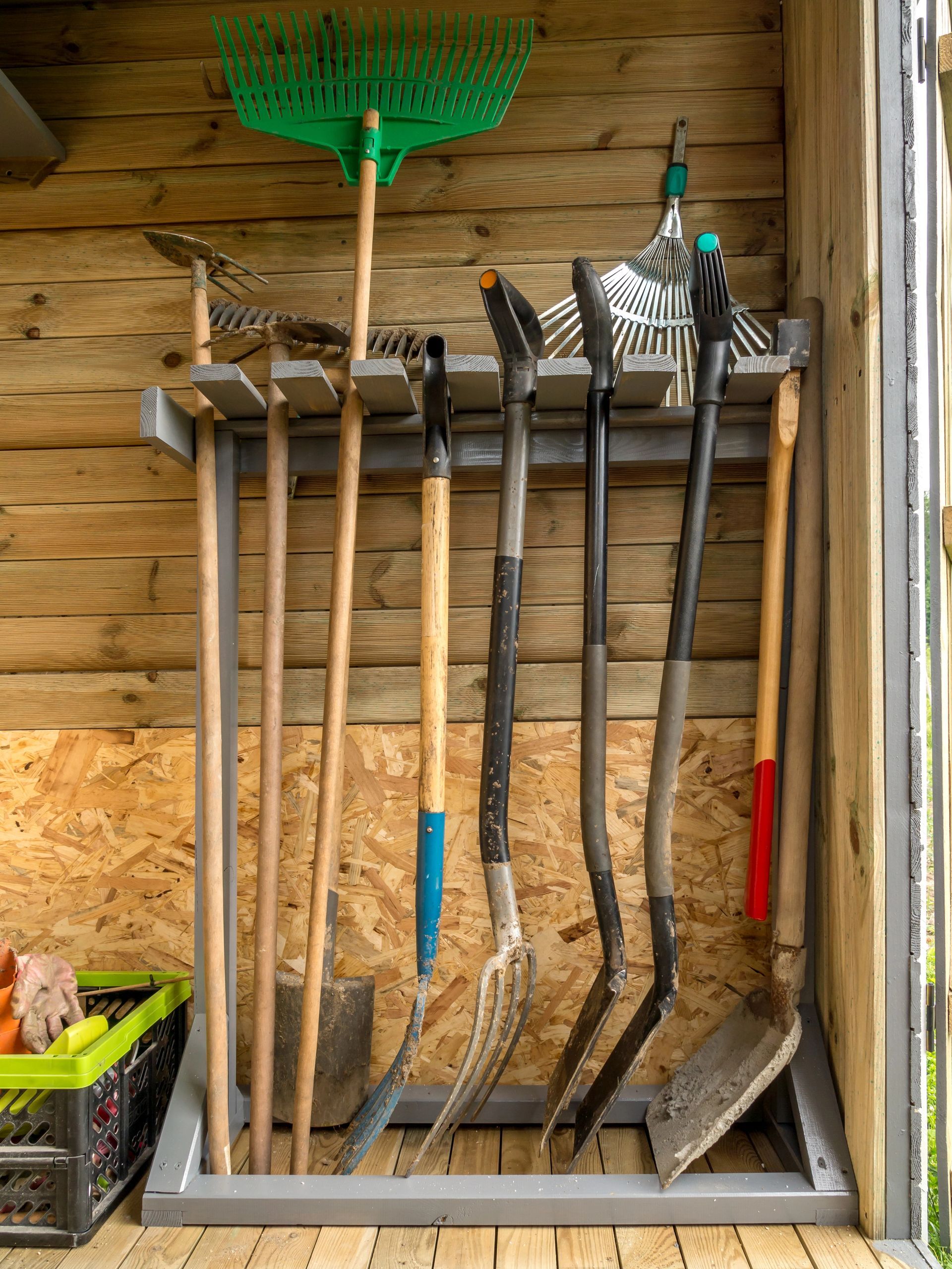 Attrezzi da giardino appesi alla parete di un capanno, tra cui rastrelli e pale con manici in legno.