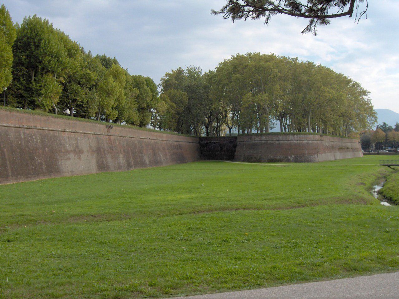 Parco erboso accanto a un lungo muro di pietra e a un terrapieno alberato sotto un cielo nuvoloso.