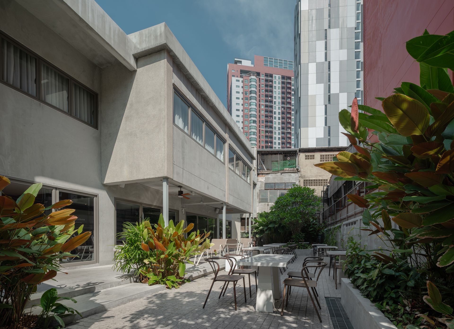 Outdoor cafe seating in a narrow courtyard between buildings, with plants and tall city towers beyond.