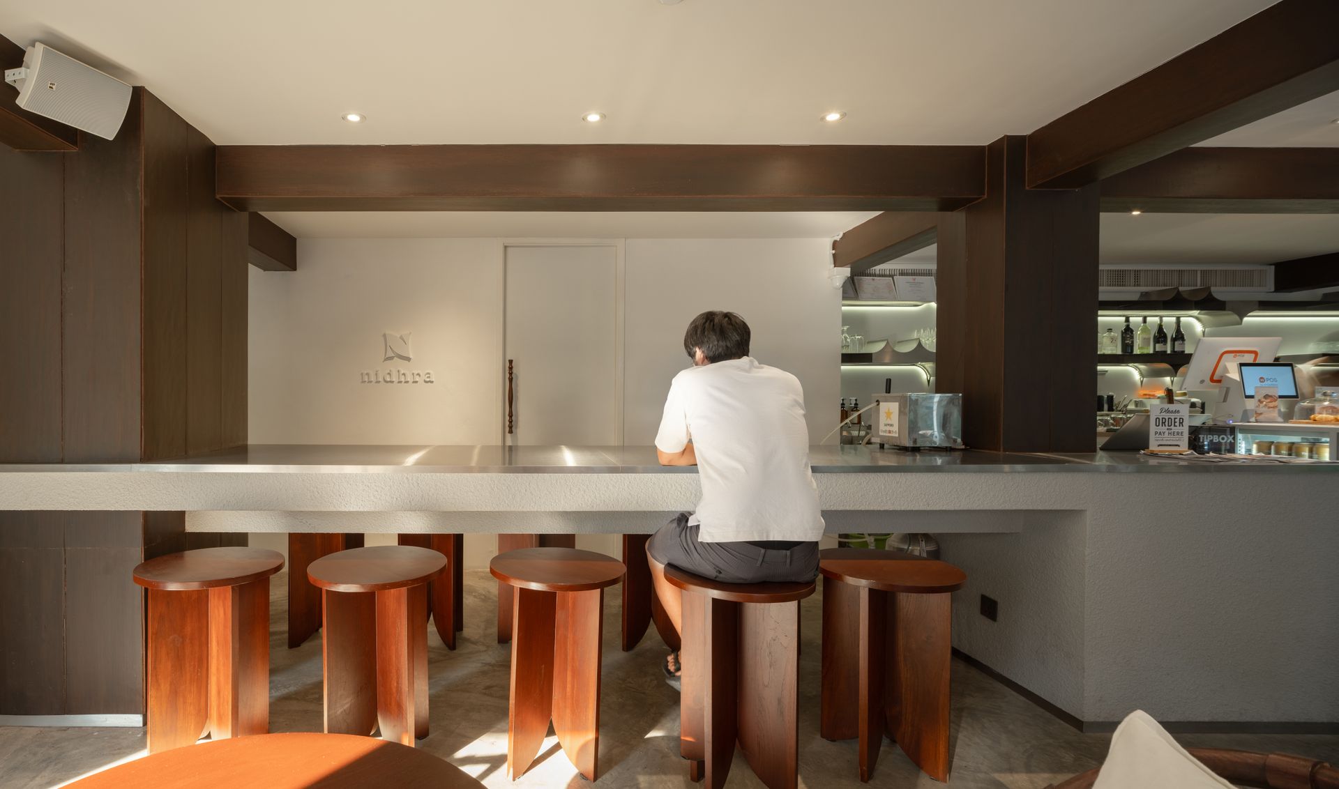 Person sitting at a wooden counter in a modern café, viewed from behind, with stools and warm lighting.