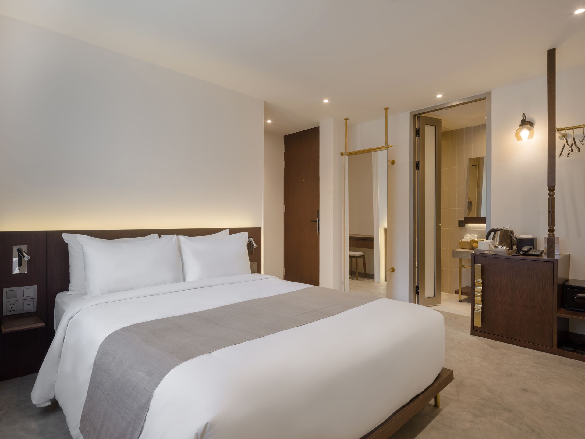 Bright hotel bedroom with a white bed, gray throw, mirrored wardrobe, and warm lighting.