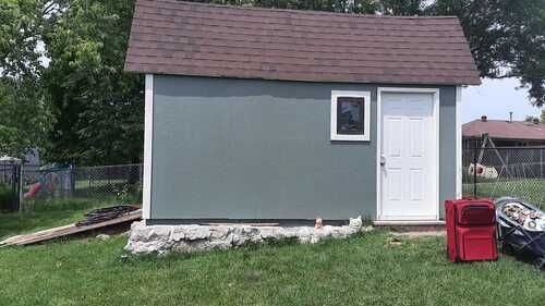 Small gray shed with a white door and window in a grassy yard, with red bins beside it.