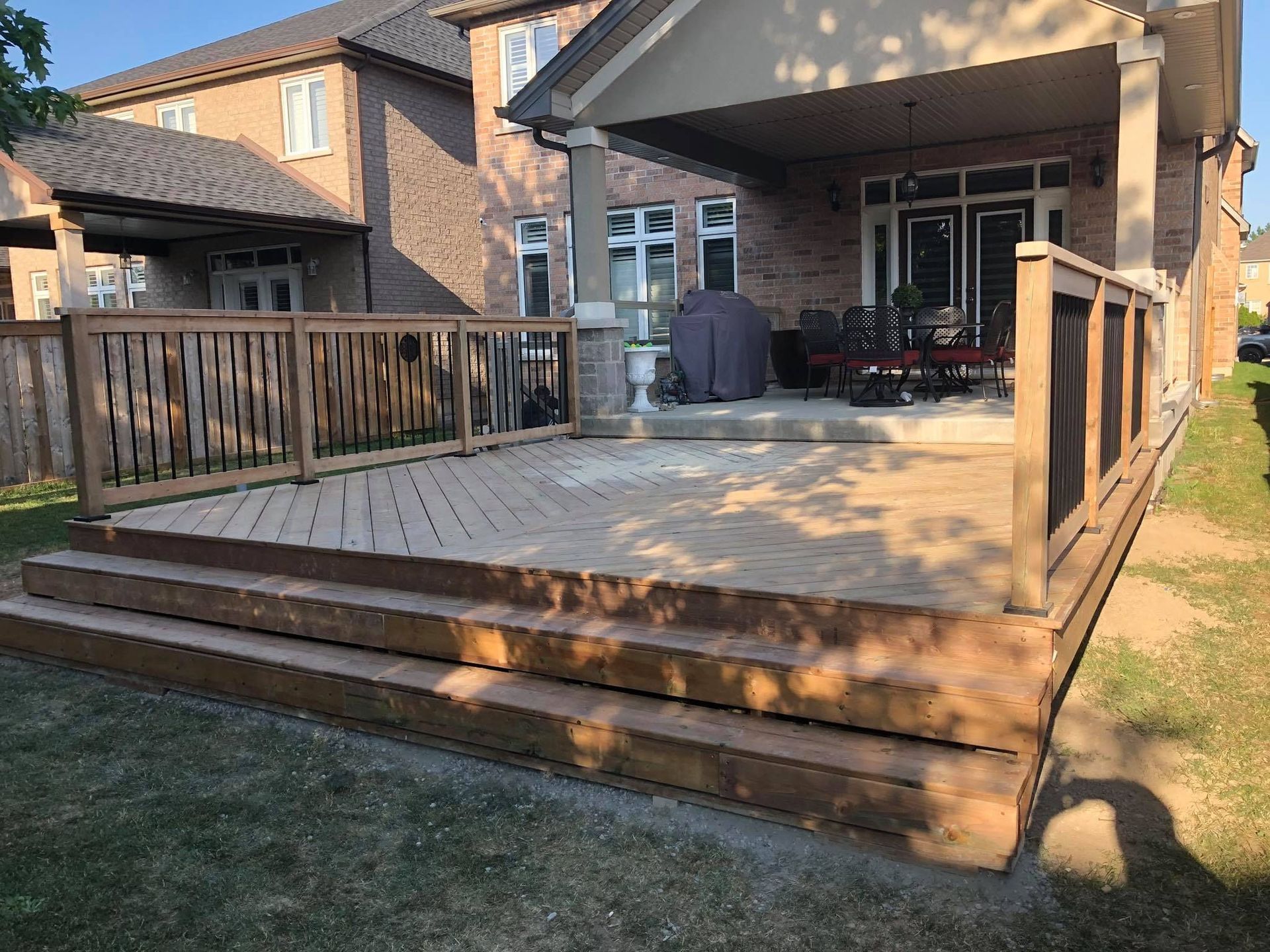 Wooden backyard deck with railing and steps, covered patio, and patio furniture beside a brick house.
