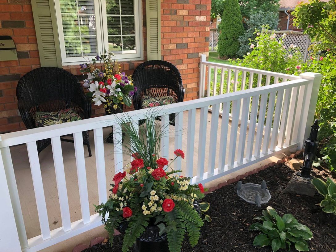 White porch railing with potted red flowers and two wicker chairs beside a brick house.