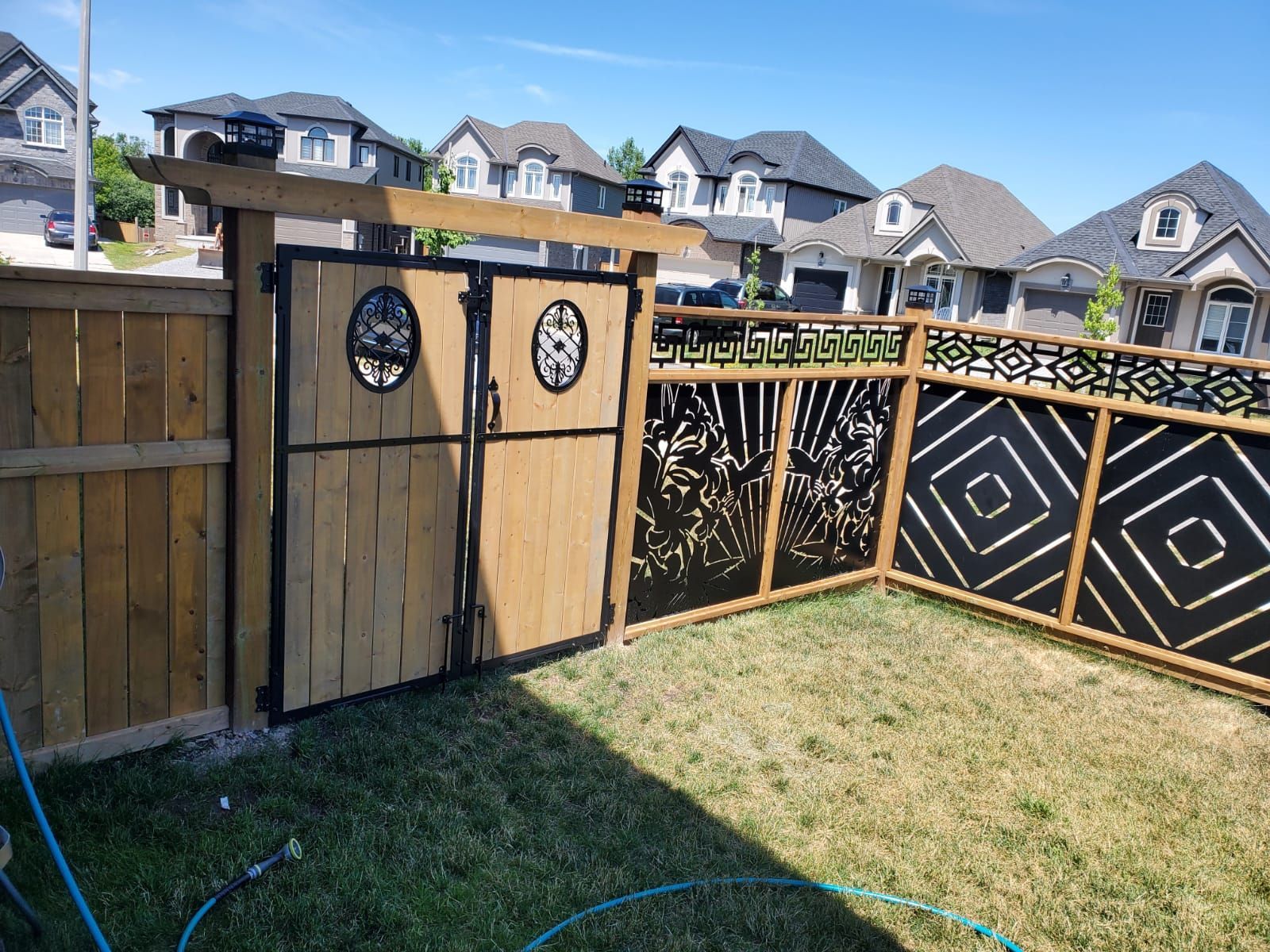 Backyard corner with wooden privacy fence, decorative black panels, and neighboring houses in the background