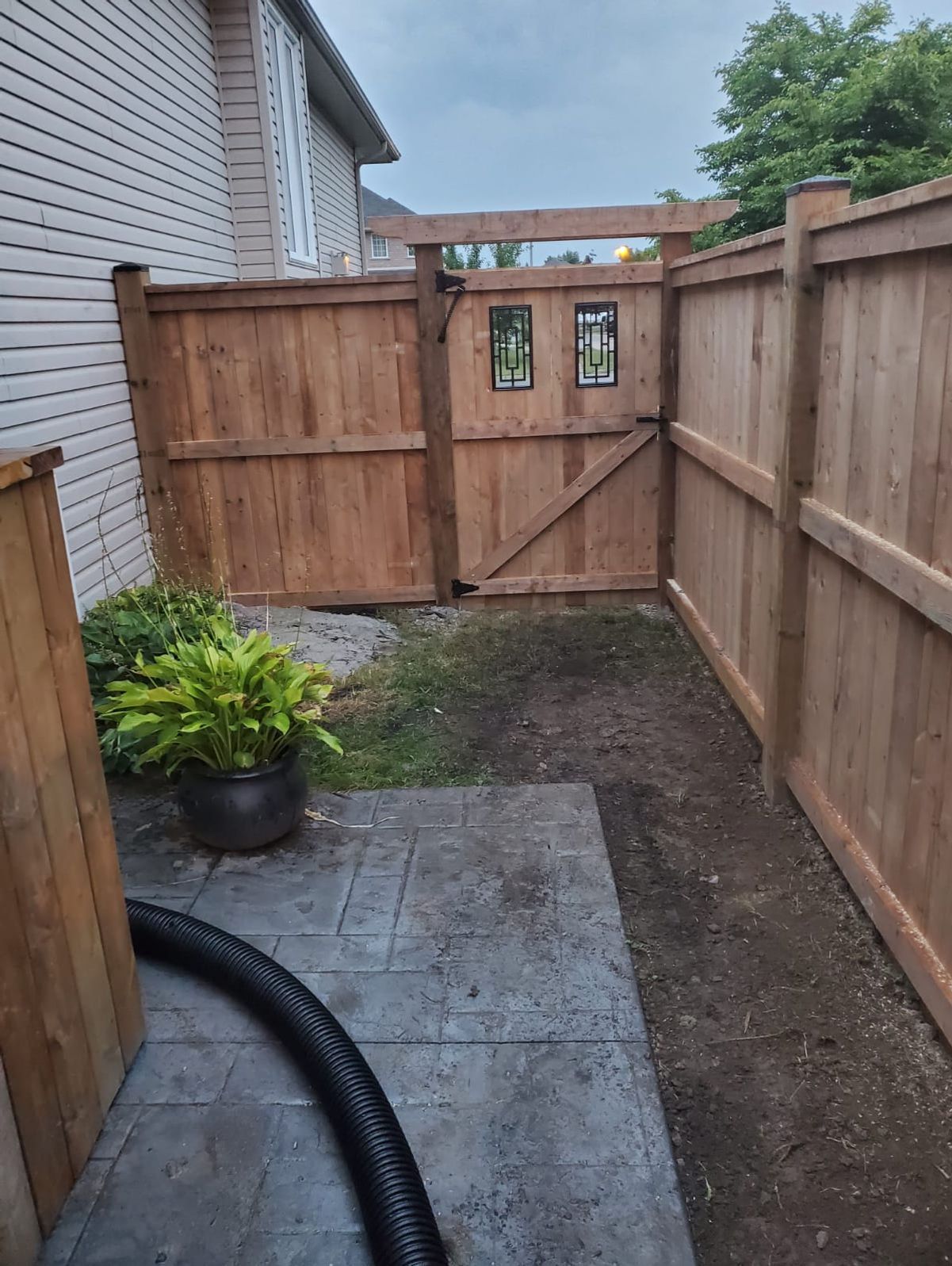 Narrow fenced backyard walkway with a wooden gate, concrete slab, and black planter beside a brick house.