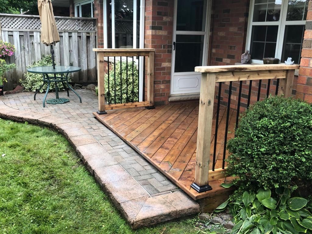 Wooden porch deck with brick steps, black railings, and a patio table beside a grassy yard.