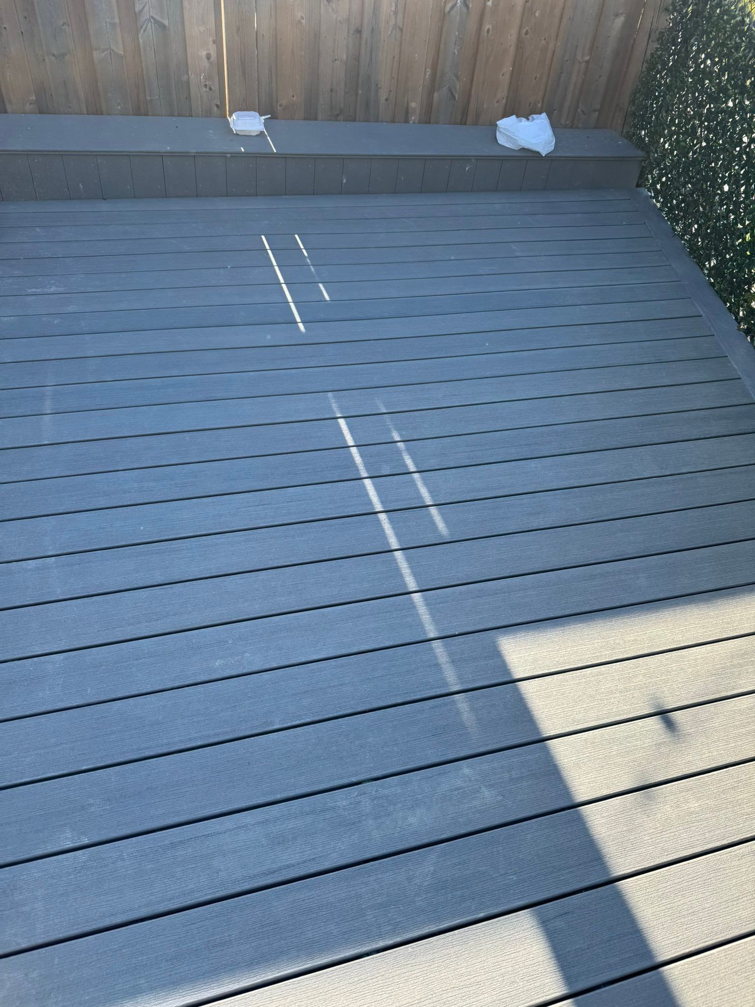 Wooden deck with railing beside a house, overlooking trees under a clear blue sky