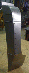 A curved, metal piece, possibly a car panel, riveted together in a workshop setting.