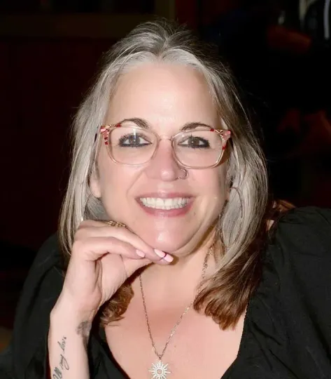 A woman with glasses and a tattoo on her wrist smiles for the camera