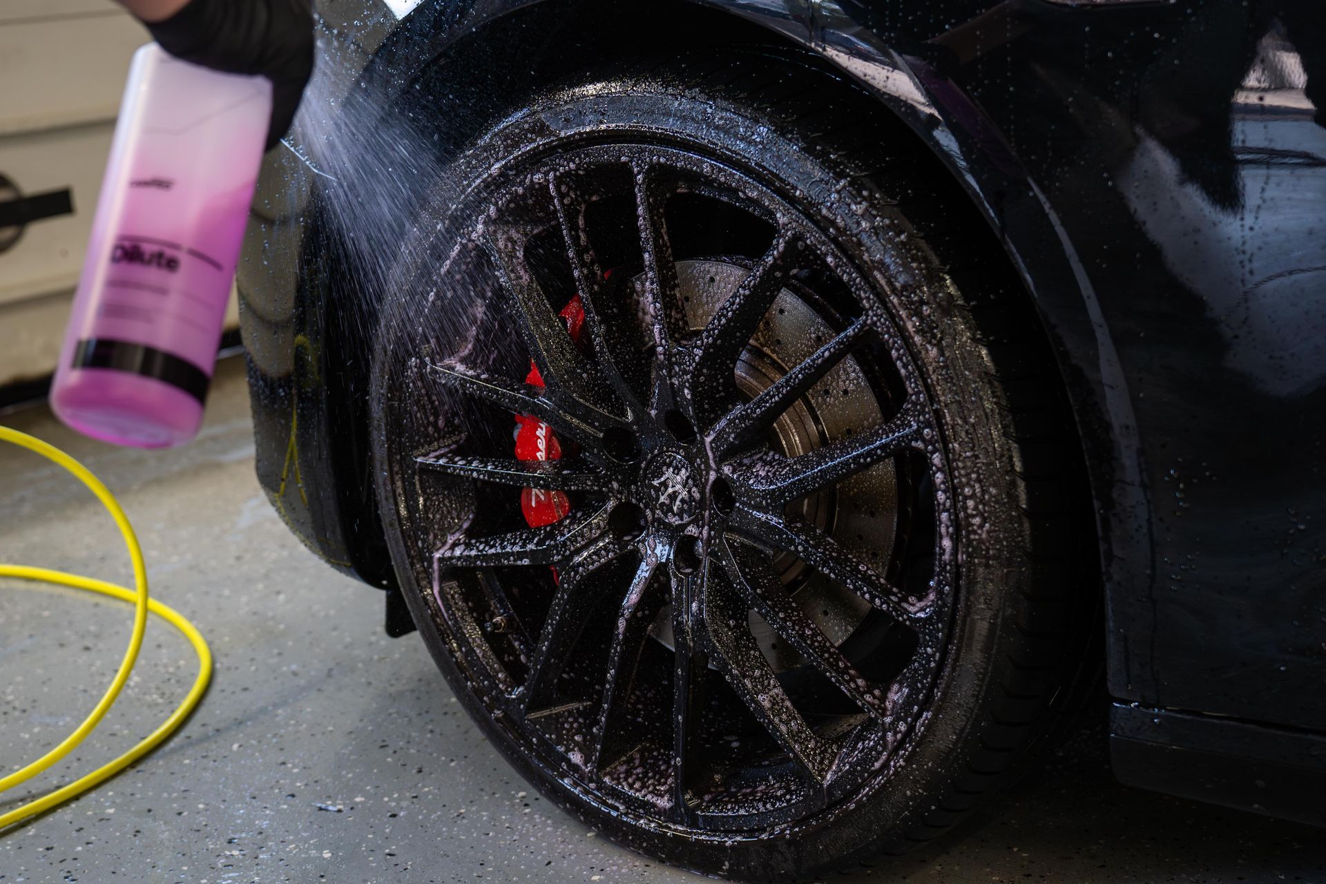Black car wheel being cleaned with a purple detailing tool, water spraying onto the rim in a garage.