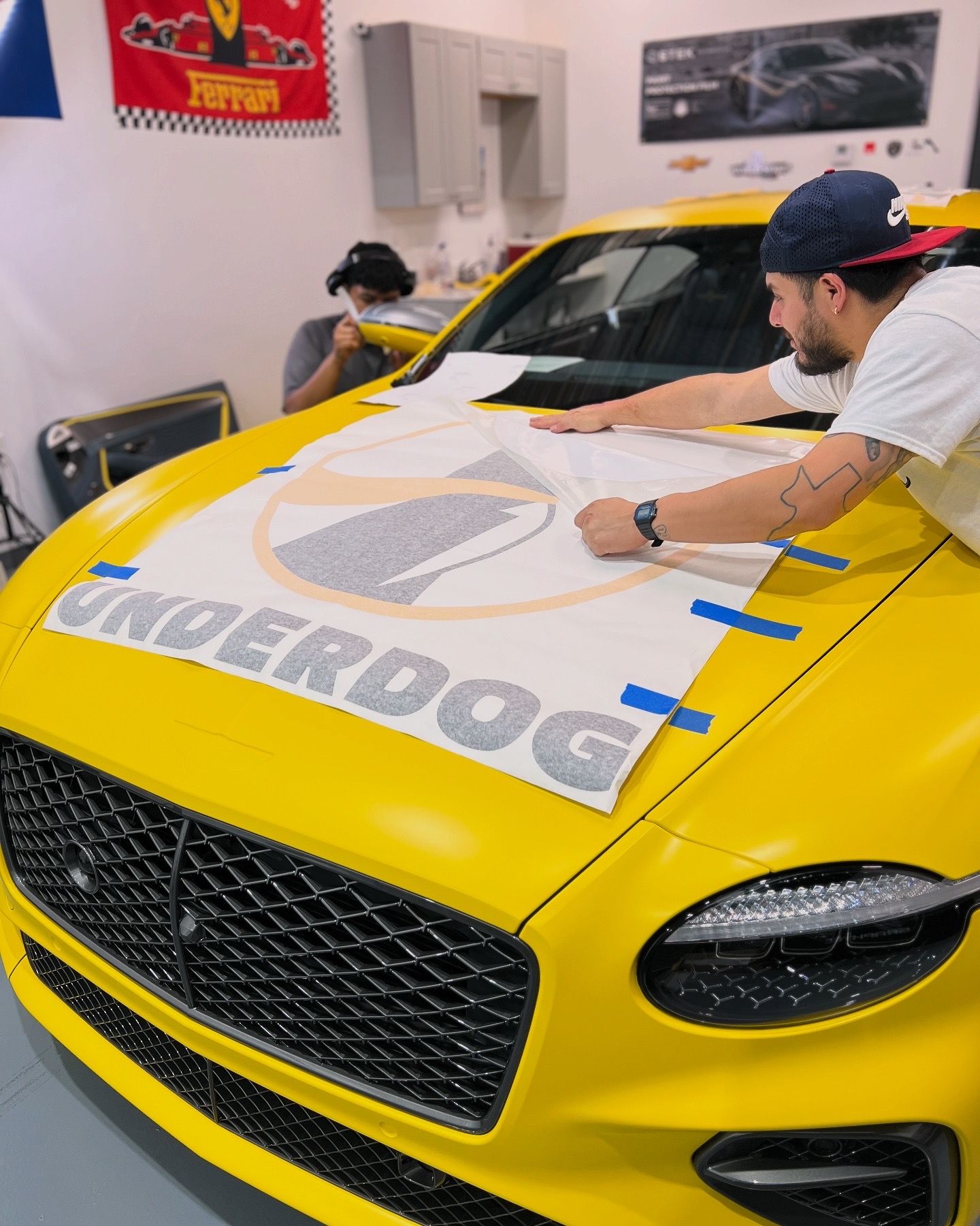 Two people applying a large decal to a yellow sports car in a workshop.
