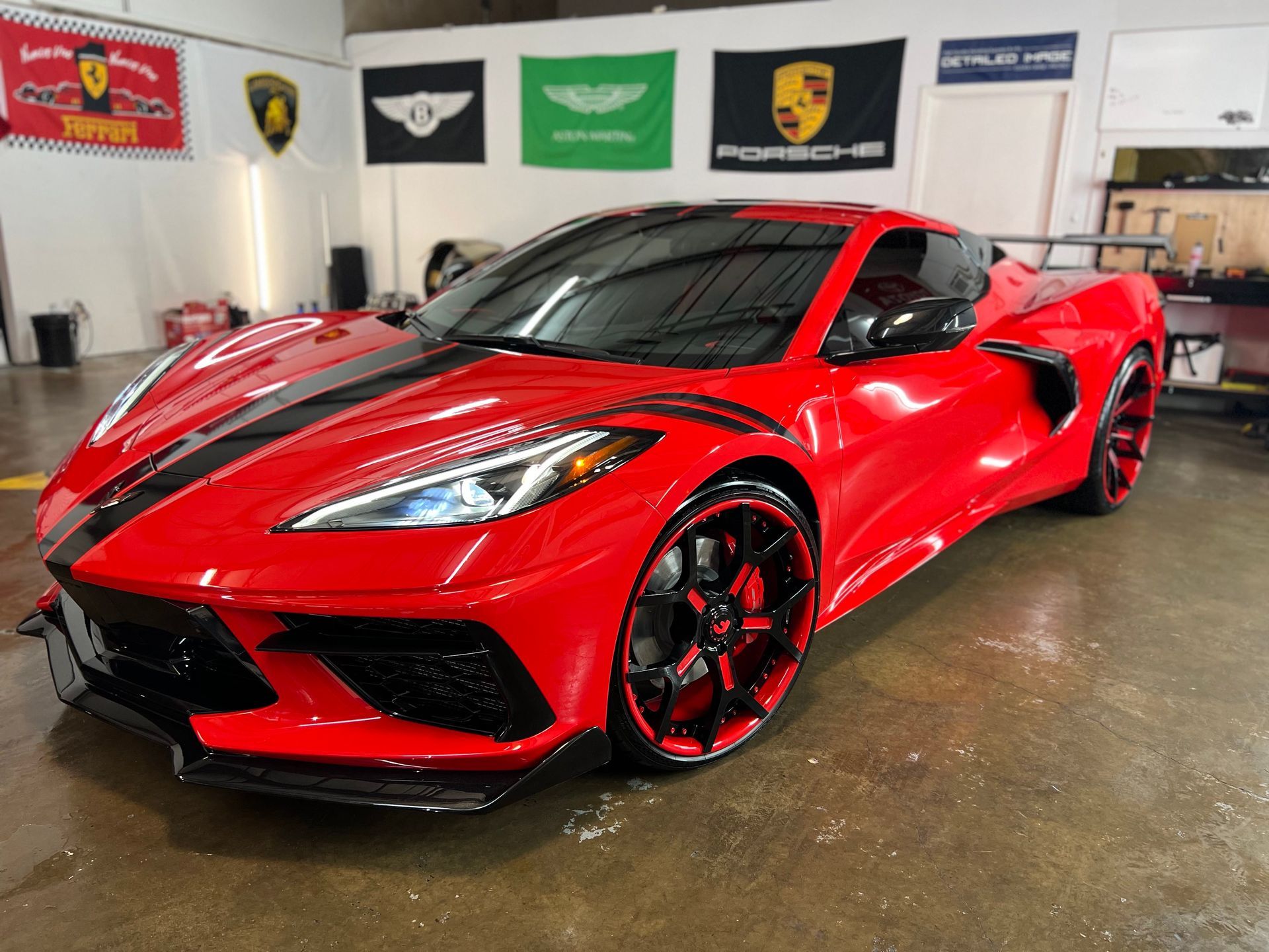 Red Corvette sports car with black racing stripes and custom rims in a garage setting with luxury car flags.