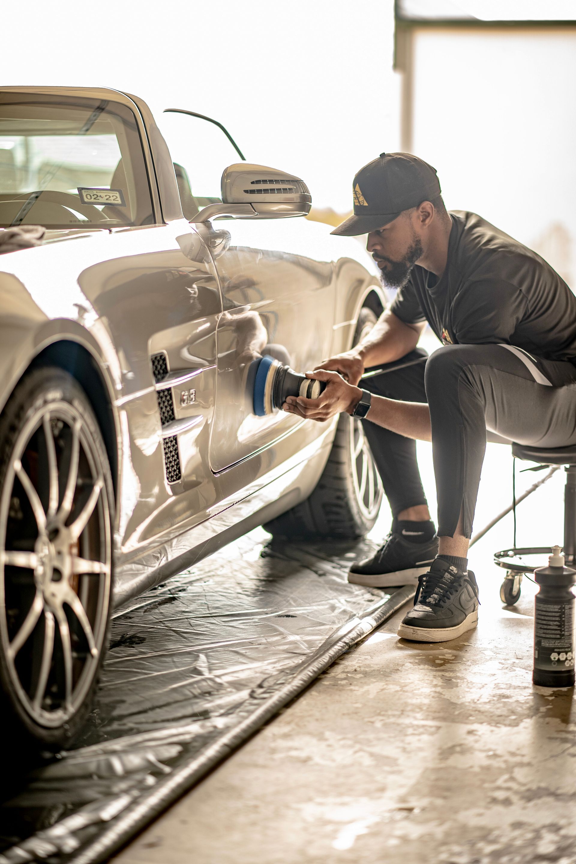 Man polishing a silver convertible car with a machine in a bright garage.