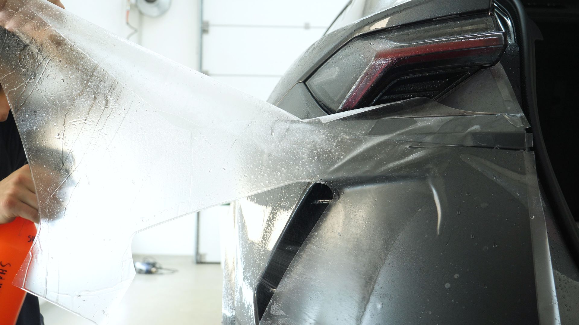 Person spray-washes the rear of a dark gray car with soapy water.