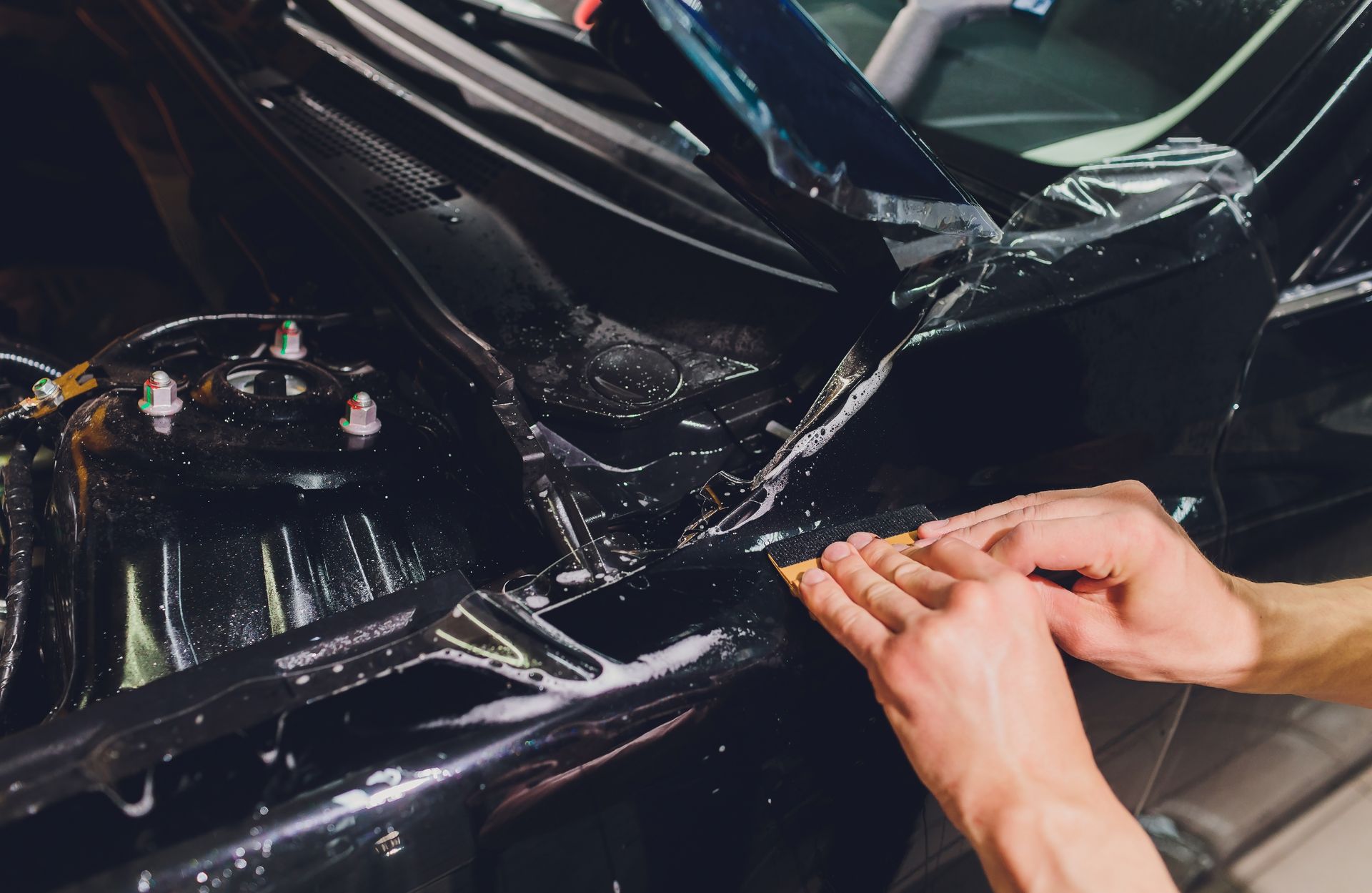 Hands working on a black car’s front body panel, applying repair or polishing tape.