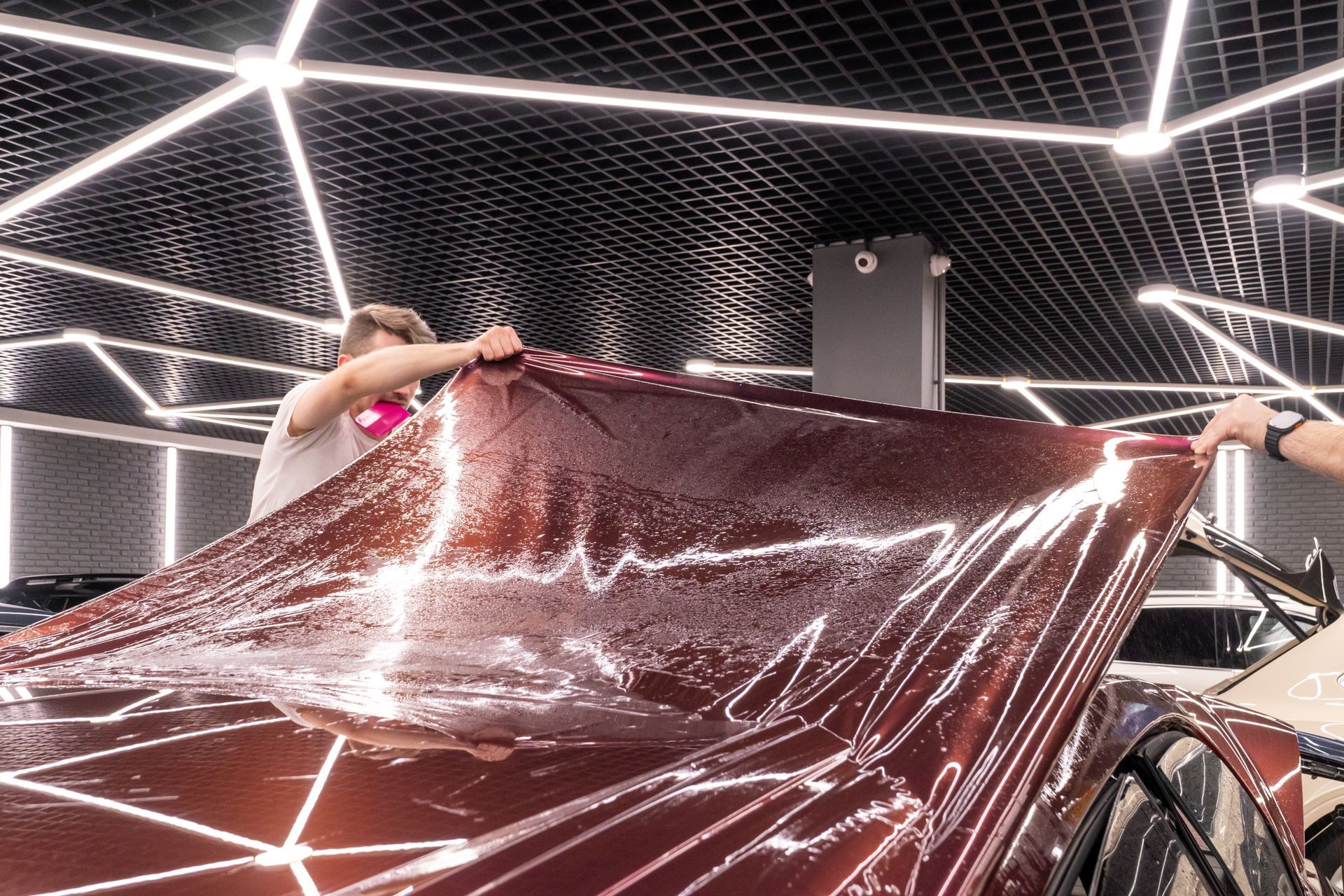 Two people holding a shiny maroon tarp under bright geometric ceiling lights in a workshop