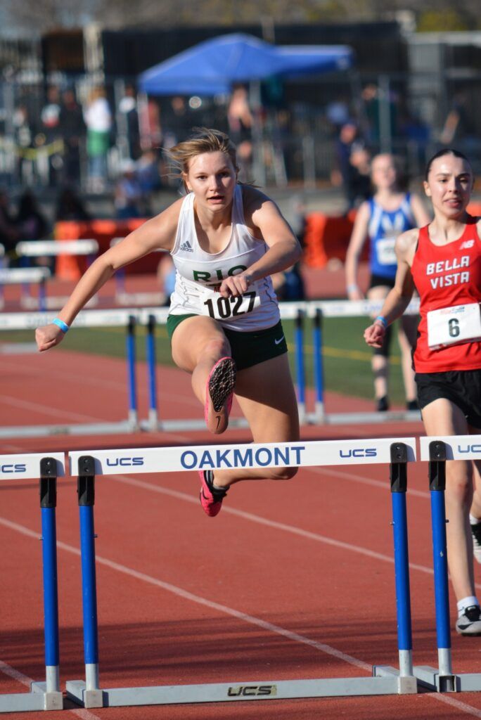 A track athlete in a white tank top clears a hurdle during a race on a sunny day.