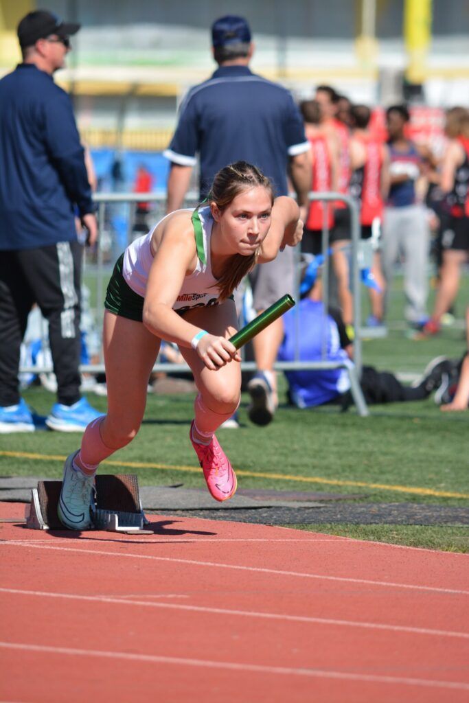 A track runner in a white tank top and dark shorts starts a race, clutching a baton while crouching at the starting blocks.