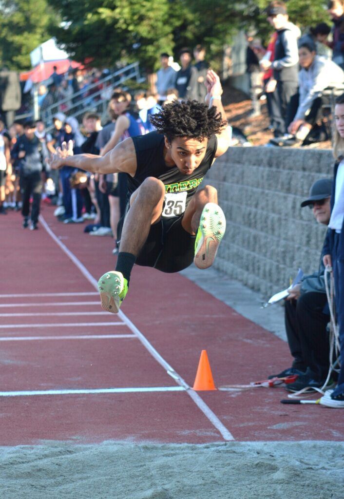 A track athlete mid-air during a long jump, wearing a black uniform and neon cleats, over a sand pit at a competition.