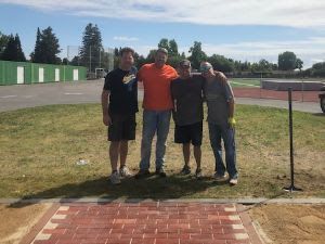 Four people standing in a row on grass in front of a brick pathway, with an athletic track and field in the background.