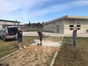 Three individuals install pavers on a sandy ground surface next to a tan building and a parked SUV.