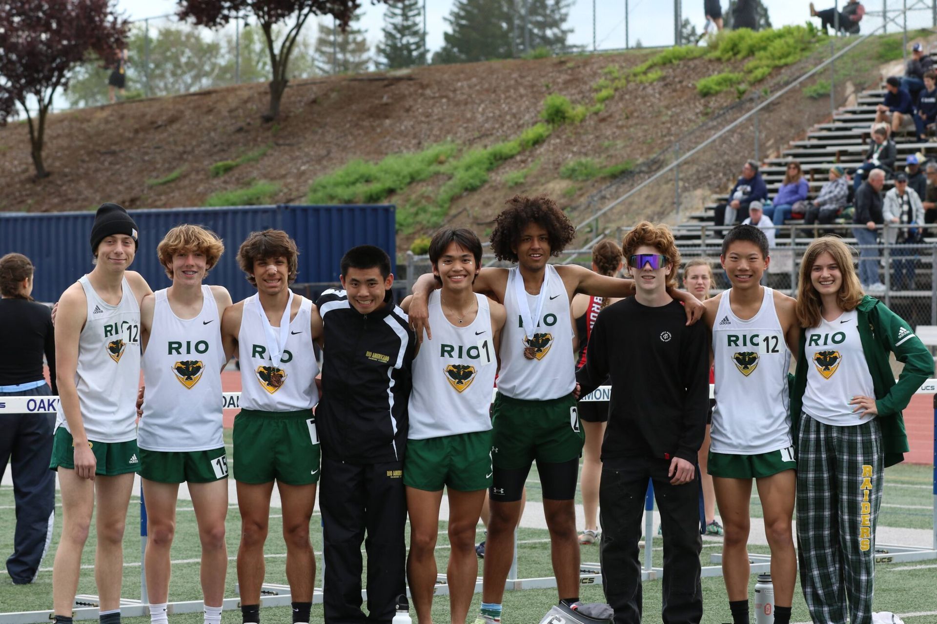 A group of ten track athletes in white Rio uniforms stand together on an outdoor track in front of stadium seating.