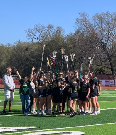 A lacrosse team gathers in a huddle on a green field, holding their sticks up together, while a man stands nearby.