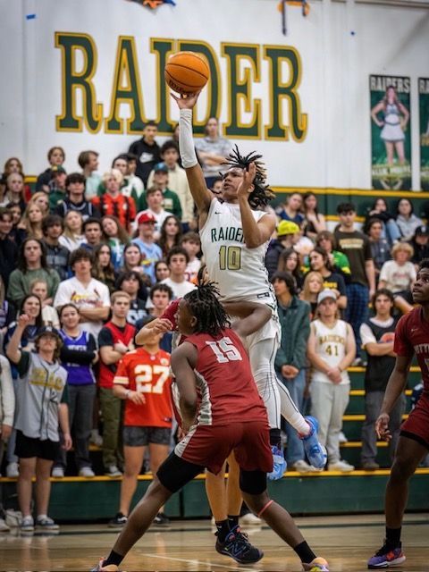 A basketball player in a white uniform jumps to shoot, defended by an opponent in red, in a crowded school gymnasium.