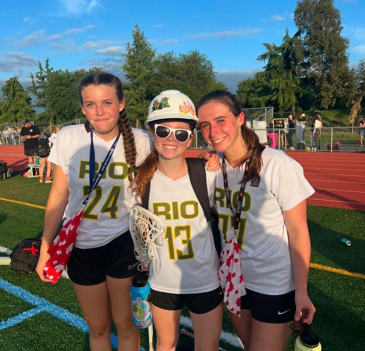 Three teammates in Rio lacrosse uniforms stand together on a sunny outdoor track field.