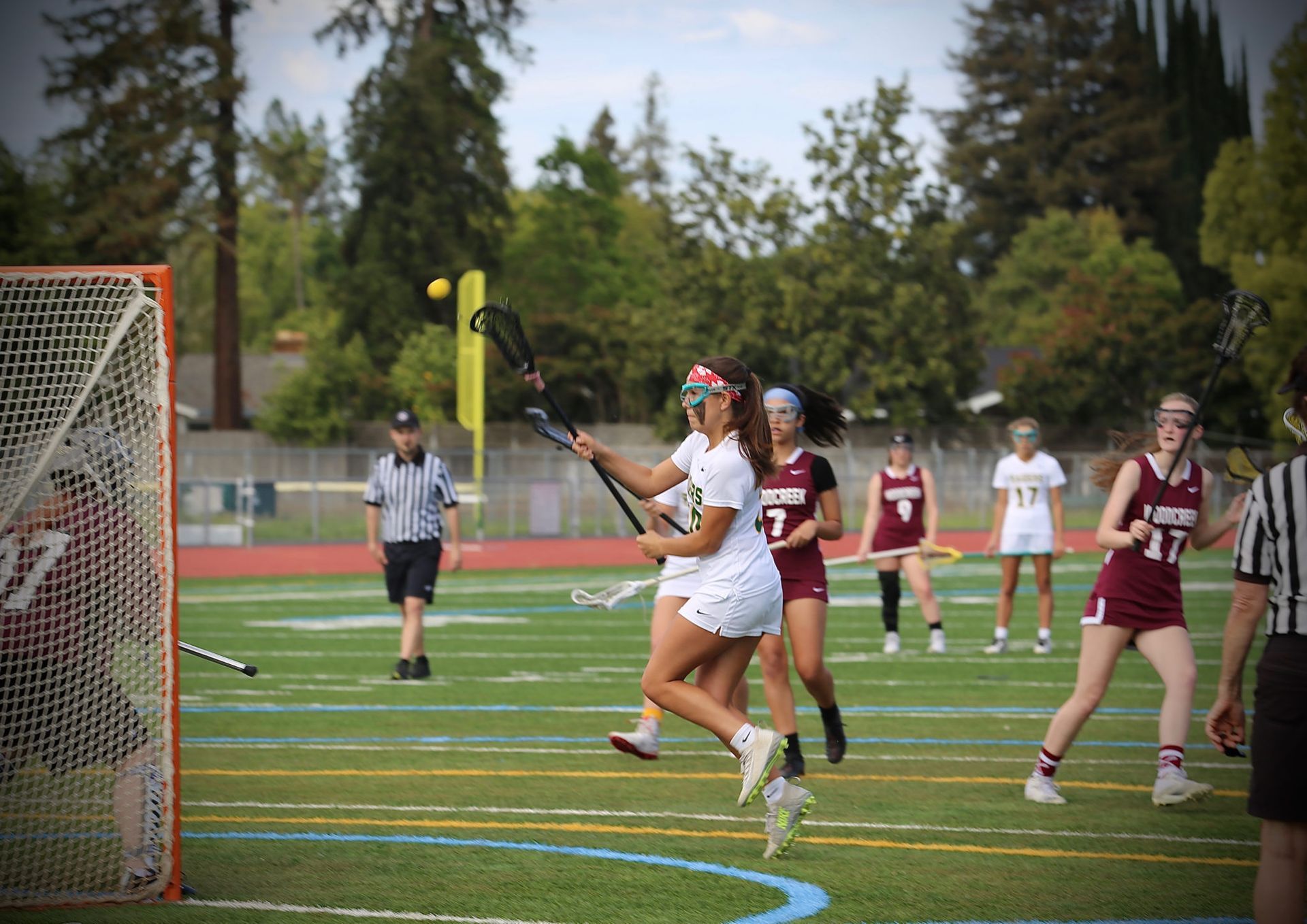 Lacrosse players in maroon and white uniforms compete on a field during a game with a referee nearby.